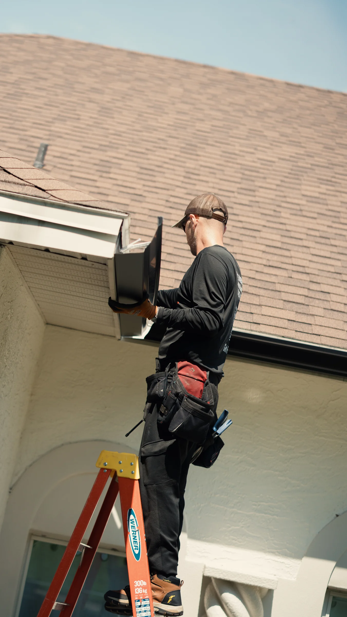 PrimeFlow field team member fitting a dark gutter section along a roof edge from a ladder